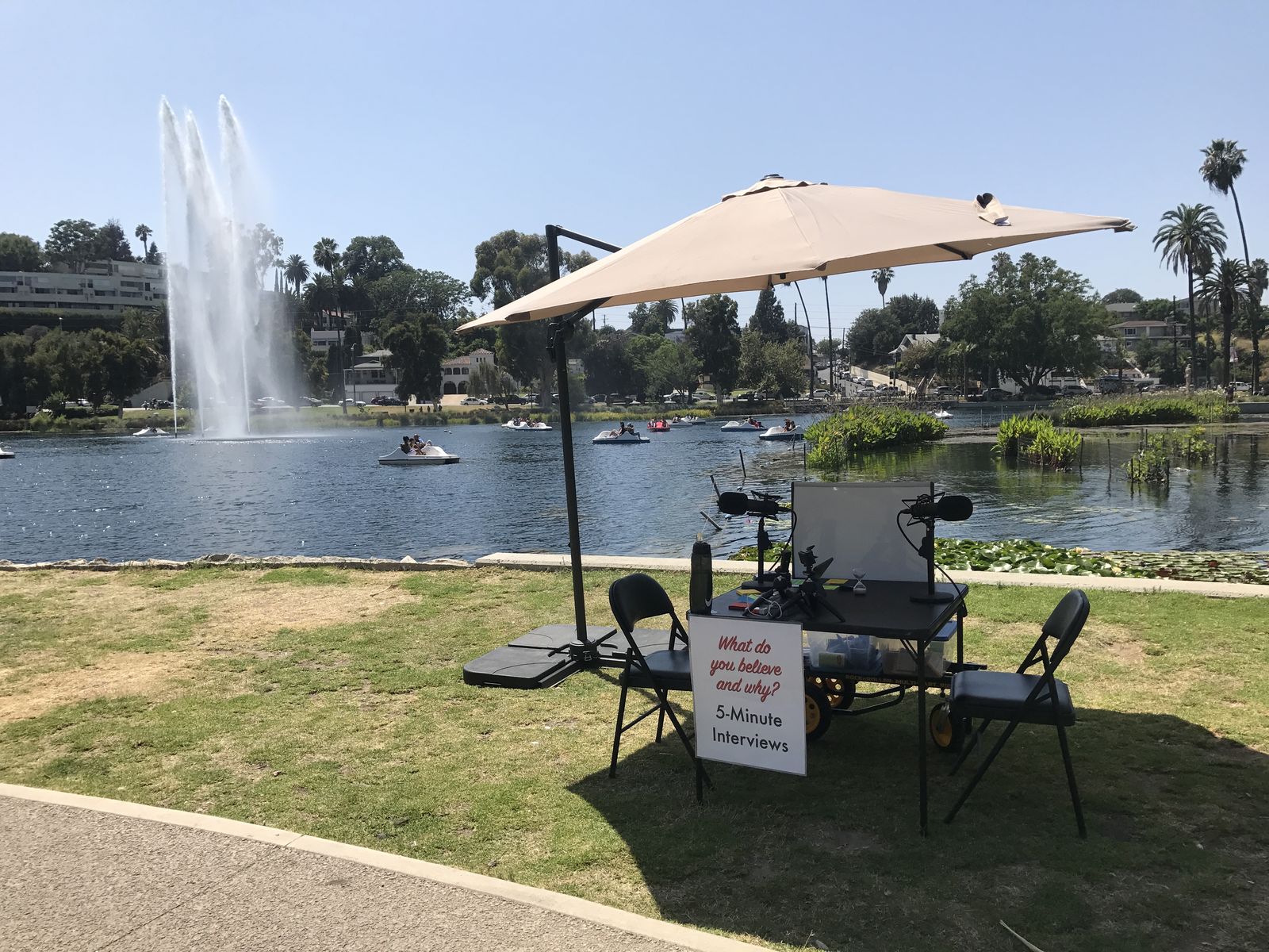A Cordial Curiosity 5-Minute Interviews table with two microphones and a patio umbrella on the grass at Echo Park Lake, with the lake fountain and pedal boats behind it